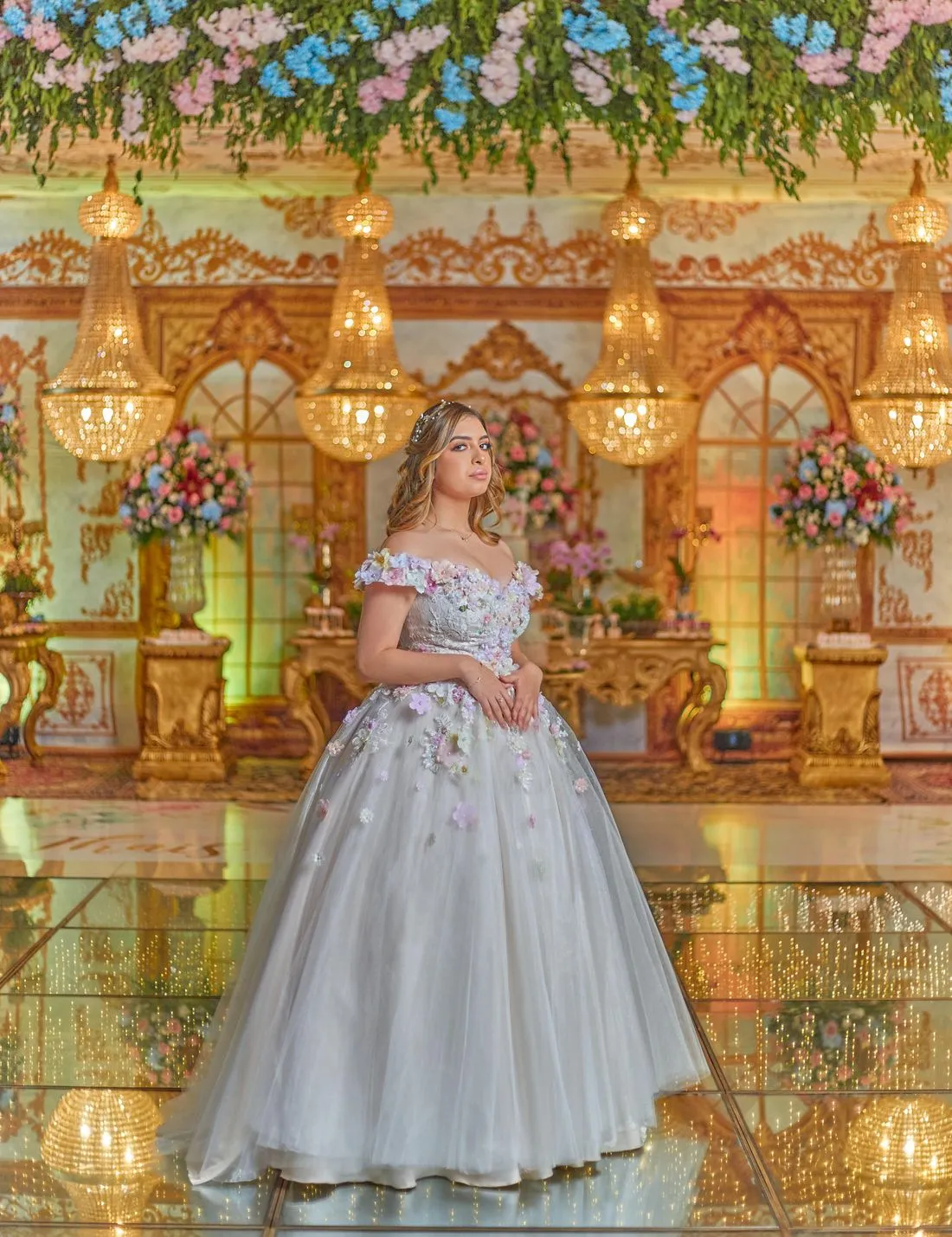 Quinceañera de pie en un salón con candelabros y arreglos de flores en el techo.