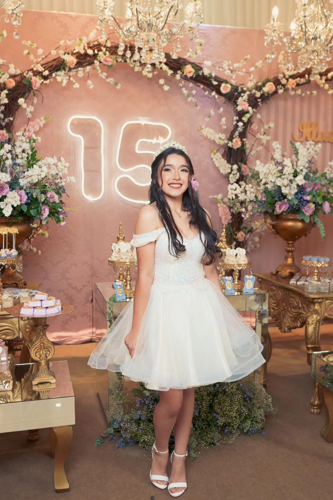 Quinceañera junto a un arco de flores con el número 15 en luces y una mesa de dulces.