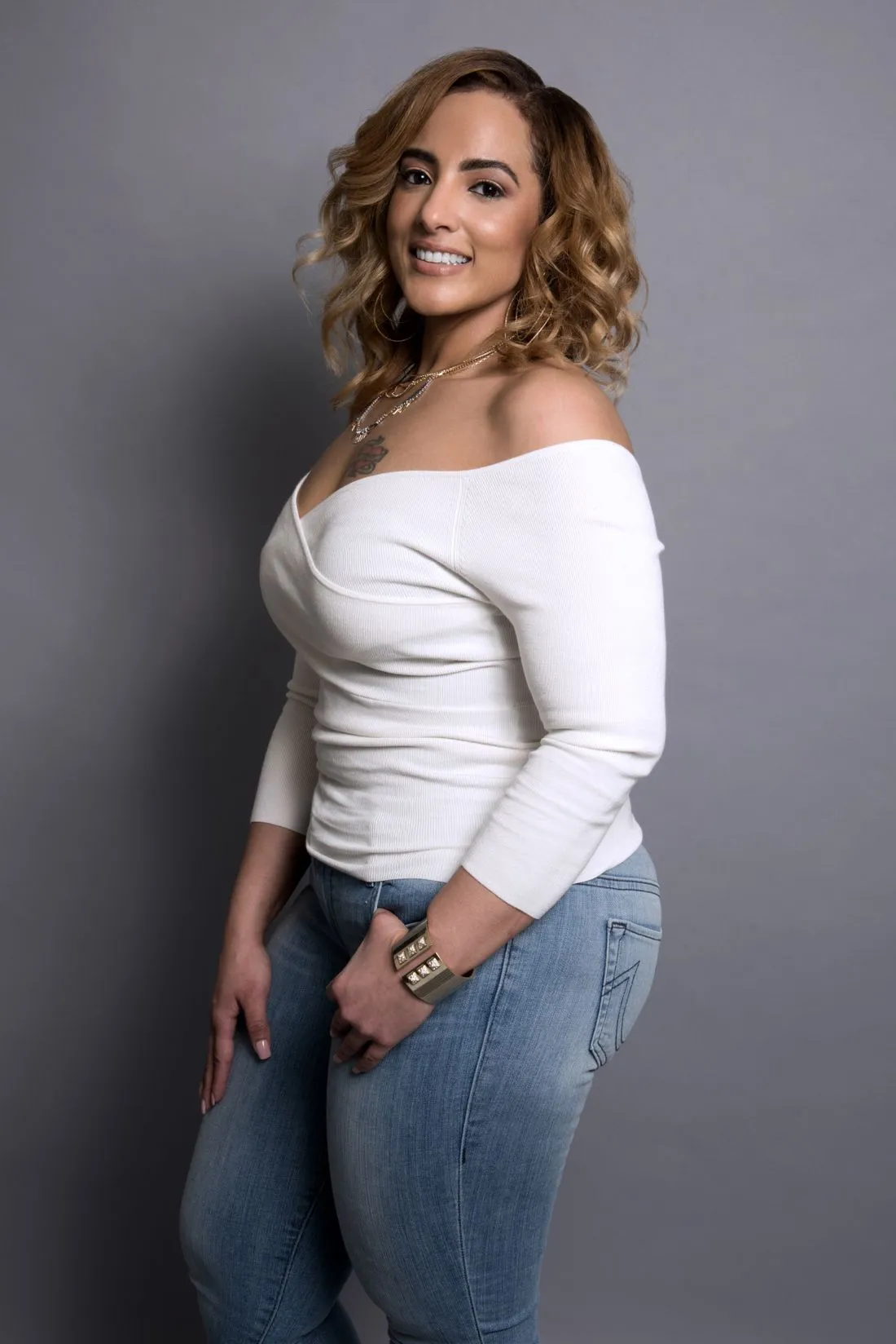 Mujer sonriente con una blusa blanca de escote amplio y jeans, frente a un fondo gris.