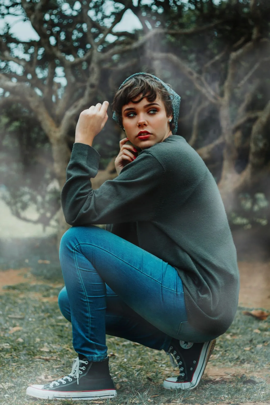 Persona agachada con jeans azules y tenis, junto a un árbol.