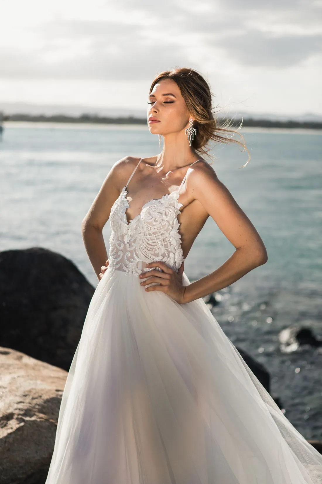 Novia de pie junto al mar con un vestido blanco de encaje en el corpiño y falda de tul.