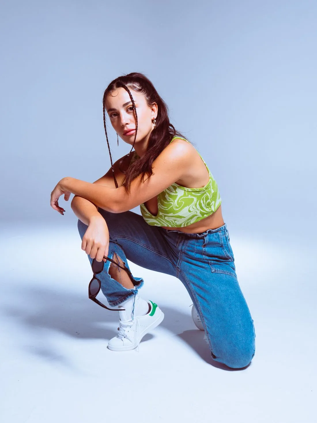 Mujer en cuclillas con jeans azules rotos, blusa verde y tenis blancos, sobre fondo celeste.