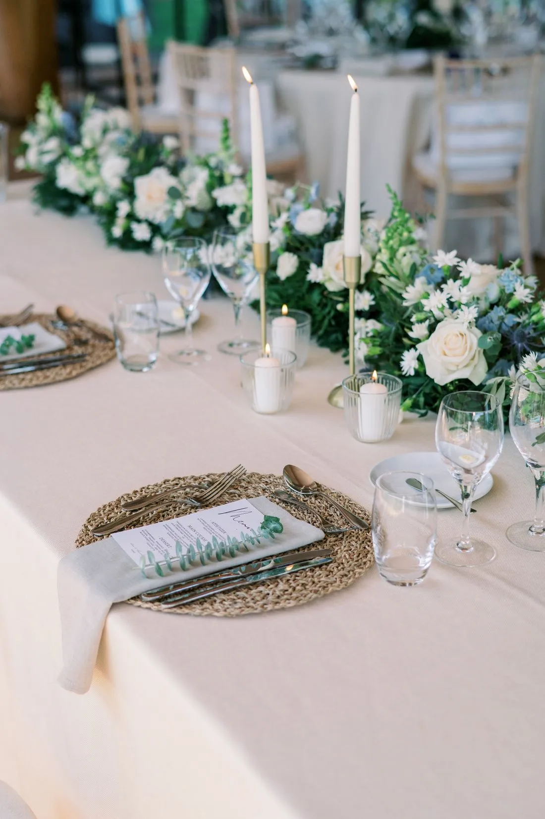 Mesa de banquete con manteles claros, velas altas y flores blancas con eucalipto.