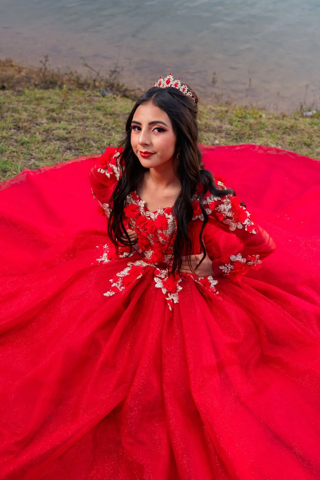 Joven con un vestido de quinceaños rojo con bordado y una tiara, sentada junto a un lago.