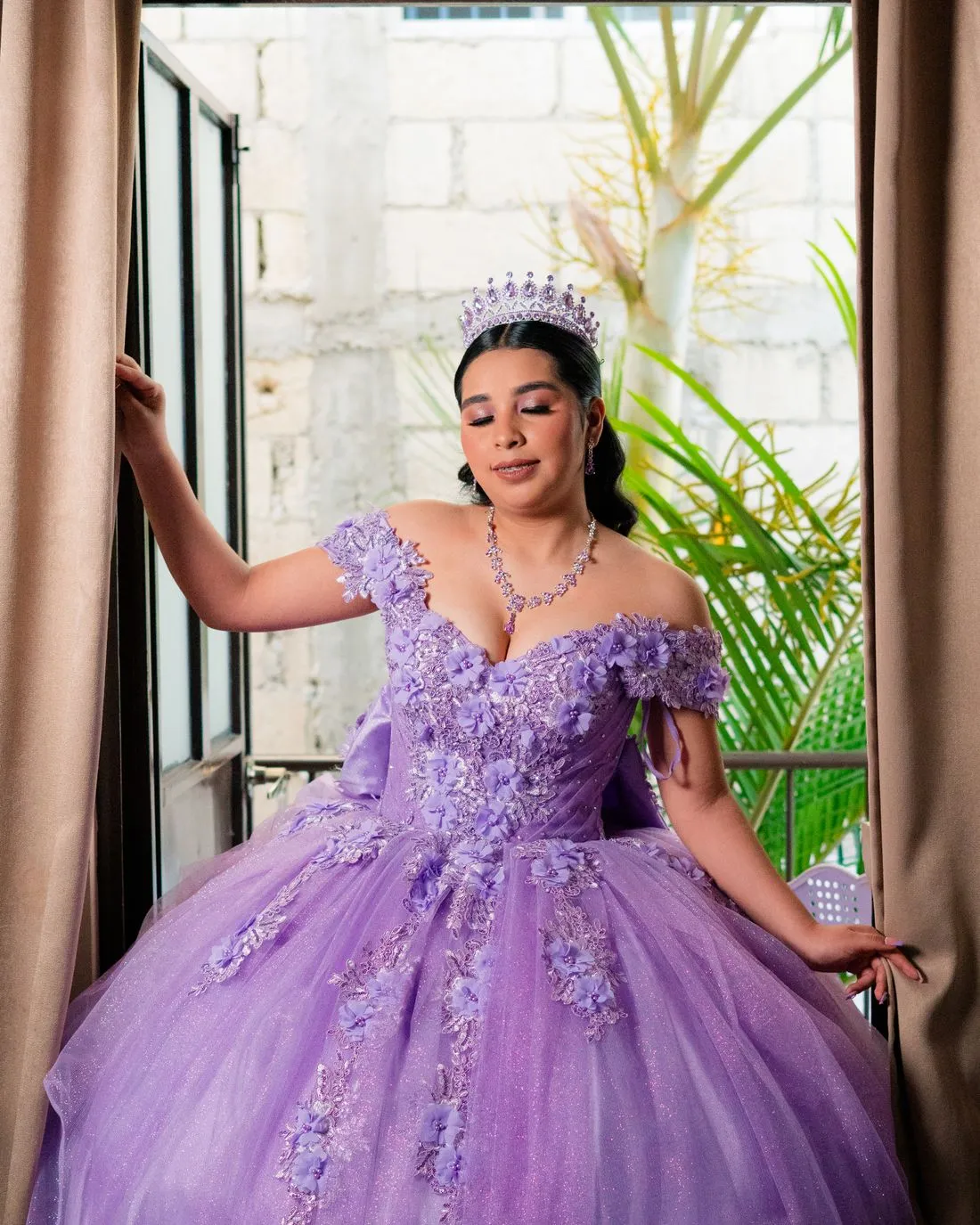 Joven con un vestido de quinceaños color lavanda con bordado de flores y una tiara, junto a una ventana.