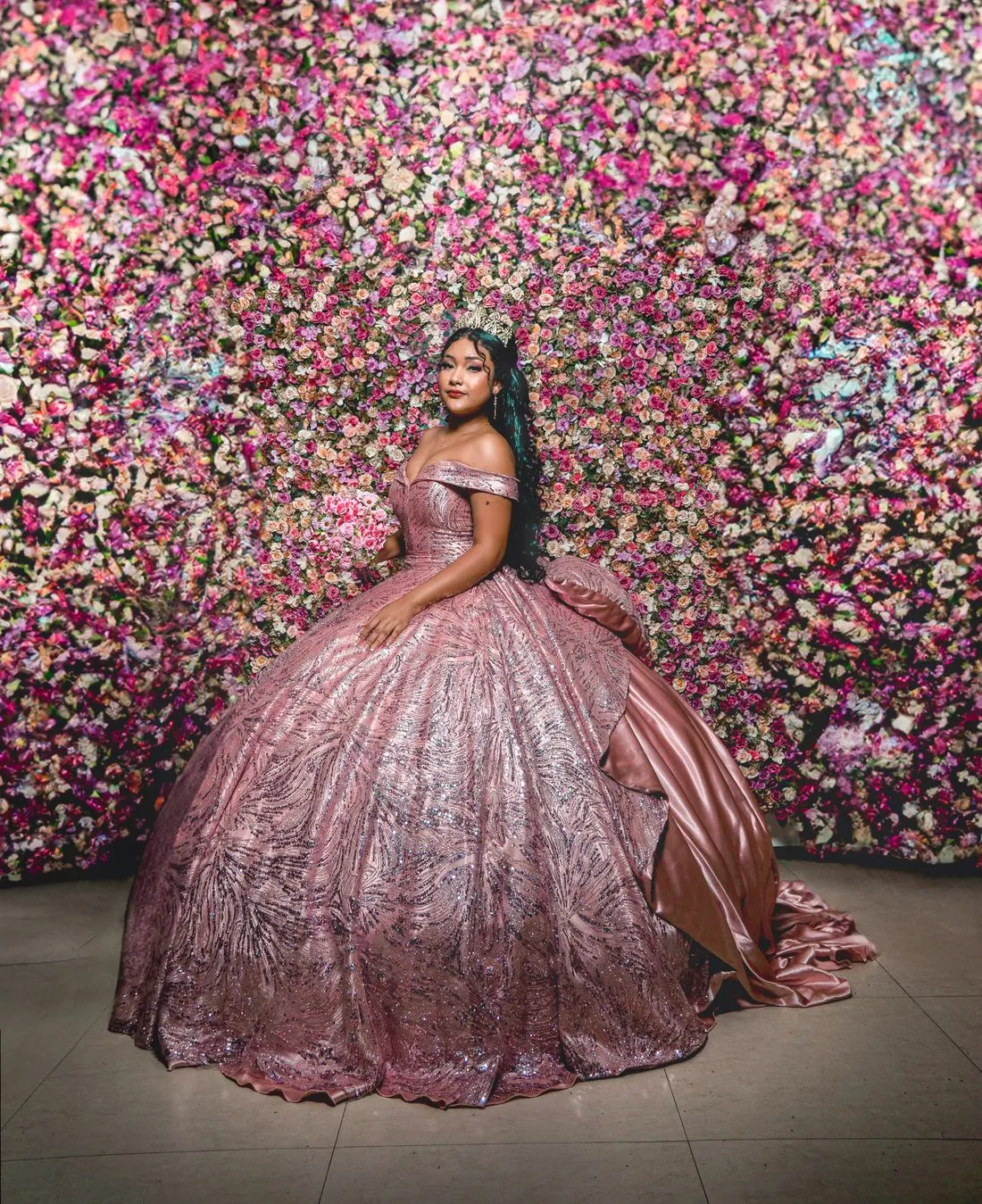 Joven con vestido rosado de lentejuelas frente a una pared cubierta de flores.