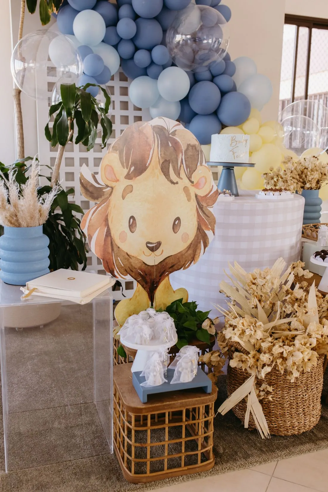 Decoración de baby shower con globos azules, un león de cartón y detalles de paja.