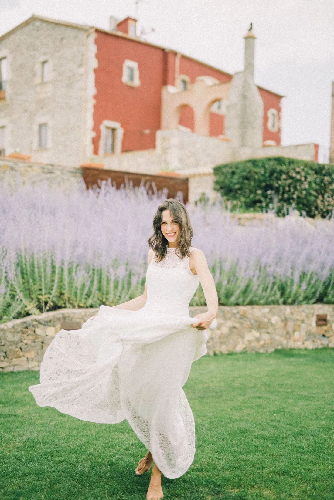 Novia en un jardín con un vestido blanco sencillo y largo, frente a una casa de piedra.