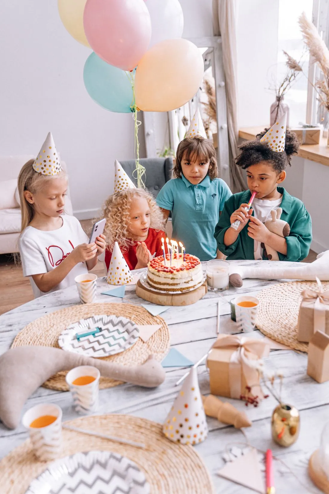 Niños con gorros de fiesta soplando las velas de una torta sobre una mesa decorada.
